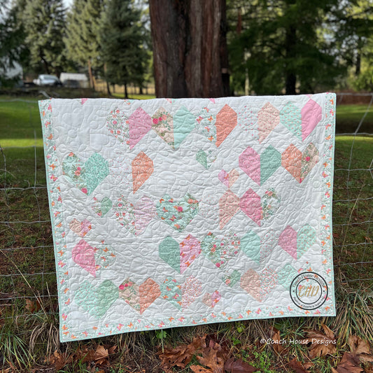 Lap quilt with two sizes of heart blocks using a different fabric for each side of the heart. Quilt is made using a Layer Cake and is shown draped over a fence.