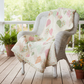 An AI image of the Kind Hearts lap quilt draped over a white wicker chair that is placed on a wooden porch, surrounded by potted plants.
